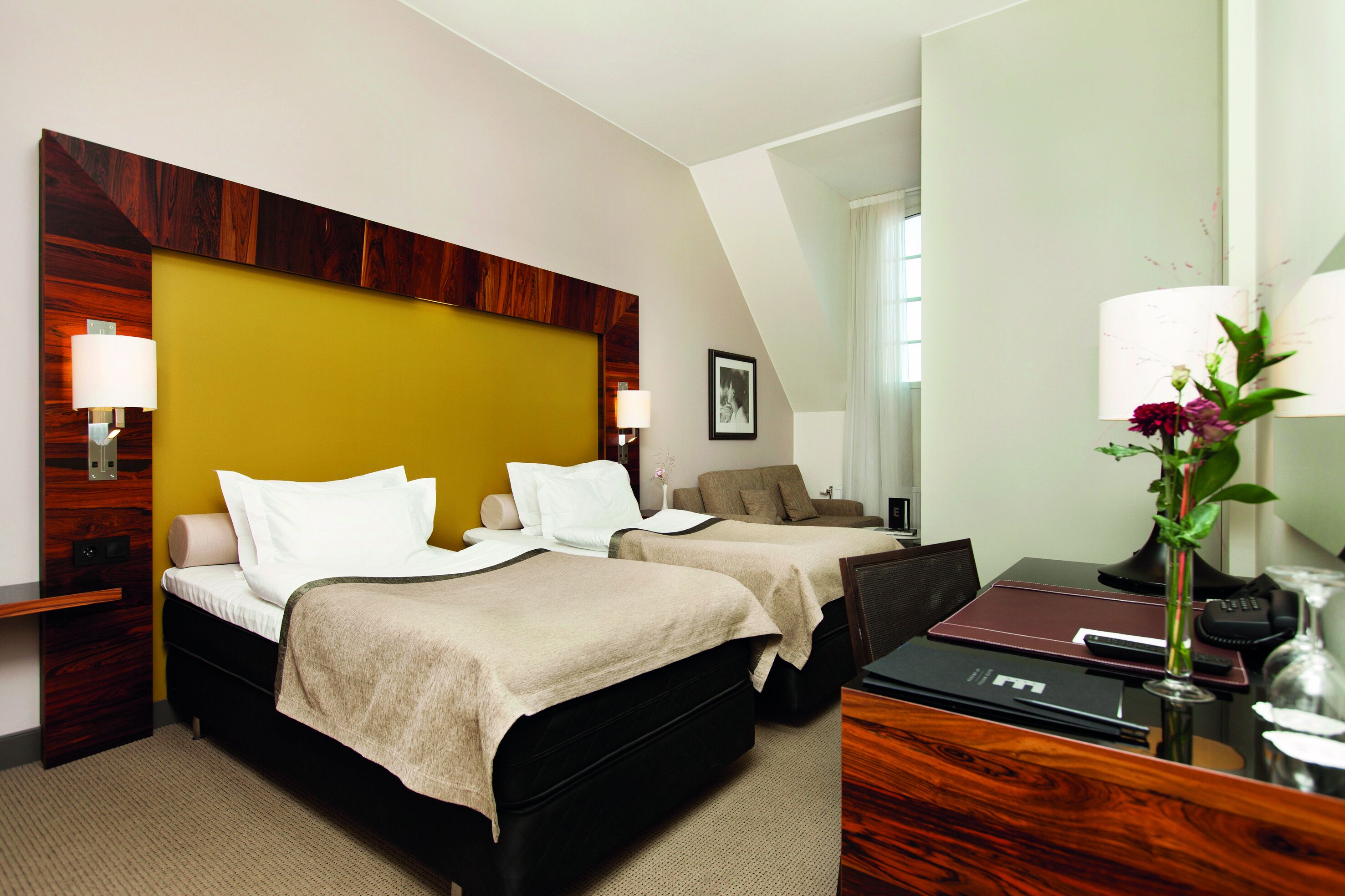 A hotel room with two beds, a desk, a chair, a lamp, and a vase with flowers.
