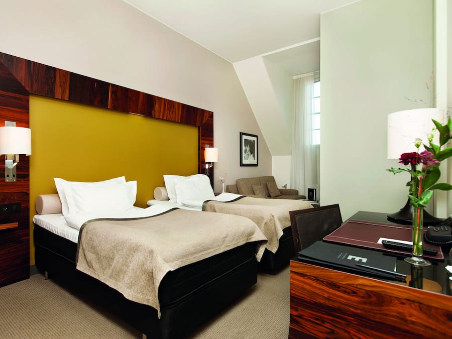 A hotel room with two beds, a desk, a chair, a lamp, and a vase with flowers.