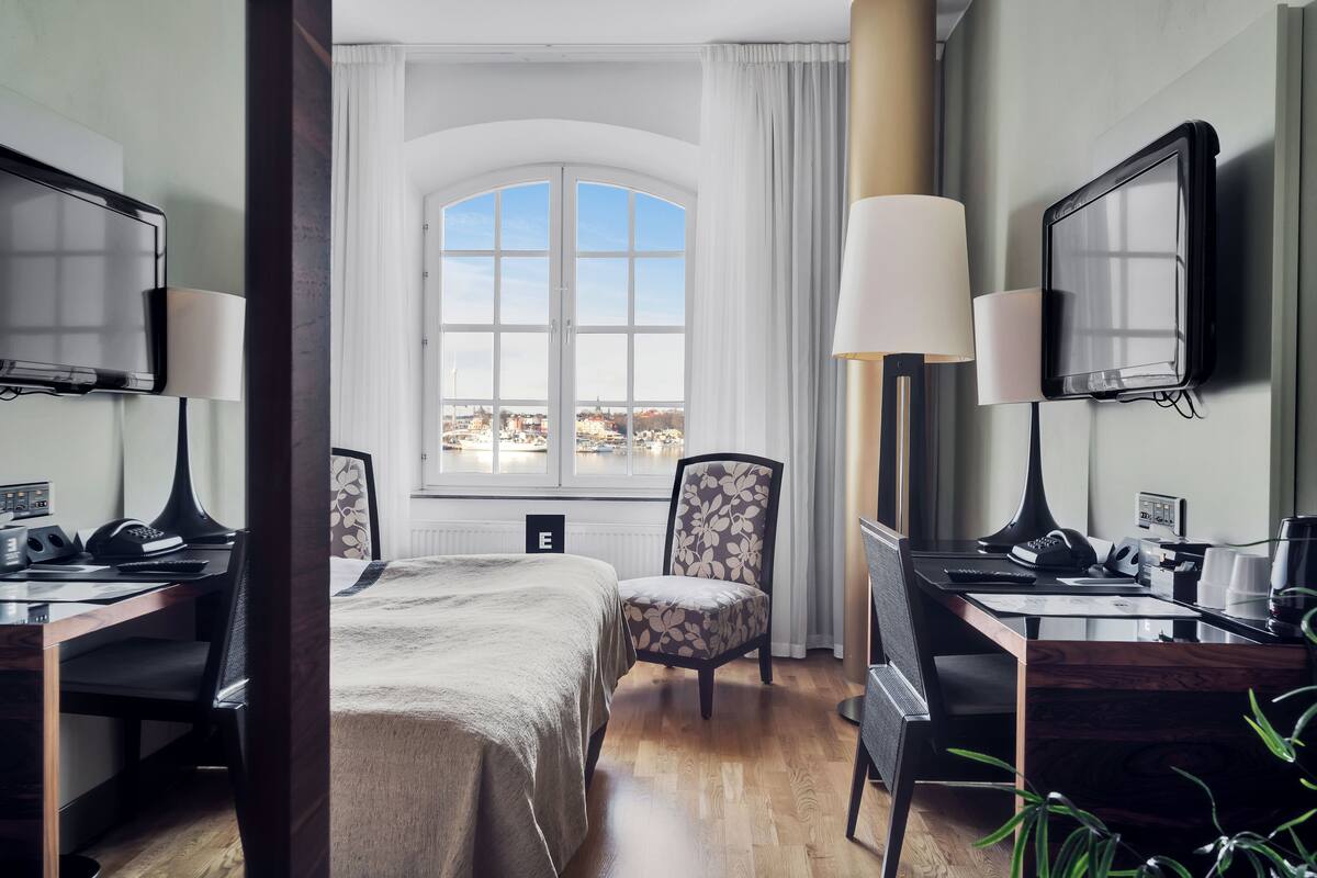 Habitación de hotel con cama, dos escritorios, una silla, televisor y ventana con vista a la ciudad.