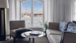 Habitación de hotel moderna con un amplio ventanal de arco que ofrece vistas al frente marítimo. Hay un sofá, una silla y una mesita redonda con libros.