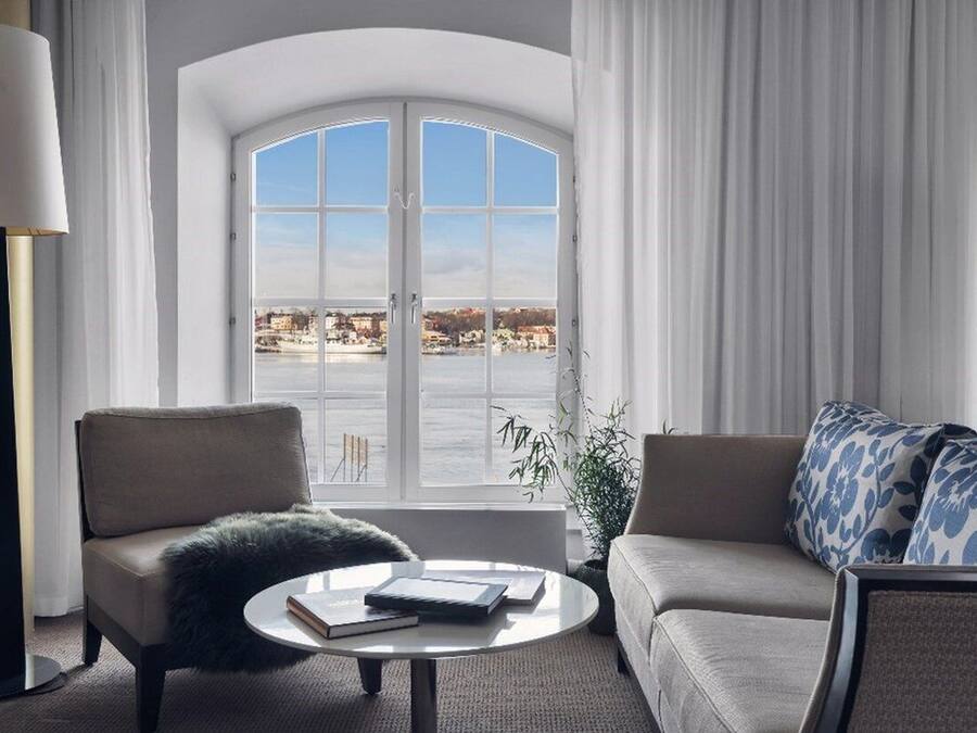 A modern hotel room with a large arched window offering a view of a waterfront. There is a sofa, a chair, and a small round table with books.