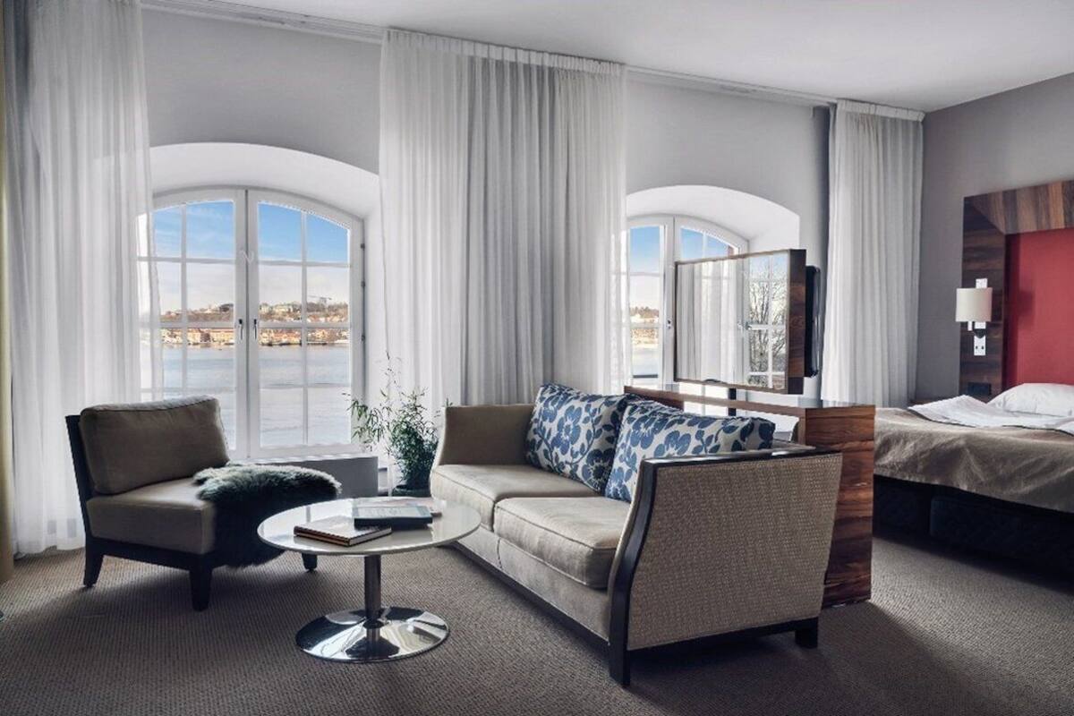 Una habitación de hotel con un ventanal grande, un sofá, una silla, una mesita y una cama.