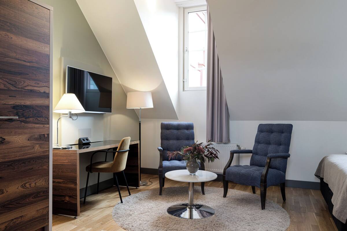 Una habitación de hotel moderna con una pared de acento de madera, un escritorio con silla, dos sillones, una mesa redonda de centro con un florero y un ventanal con cortinas.
