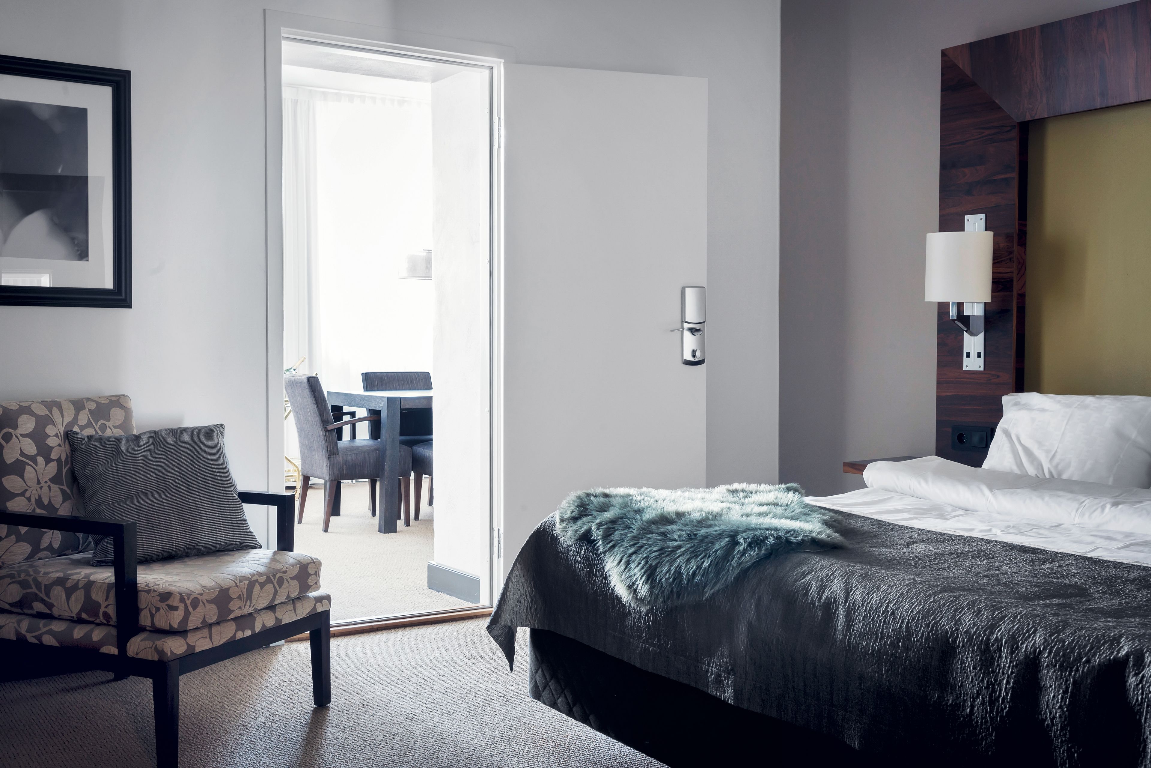 A hotel room with a bed, a chair, a desk, and a door leading to a dining area.