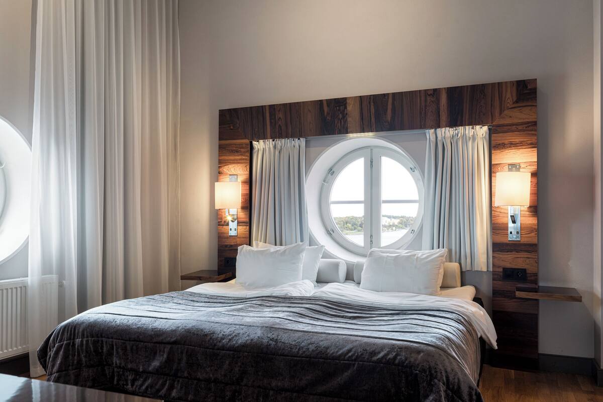 Una habitación de hotel moderna con una cama grande, un ventanal redondo y paneles de madera en las paredes.
