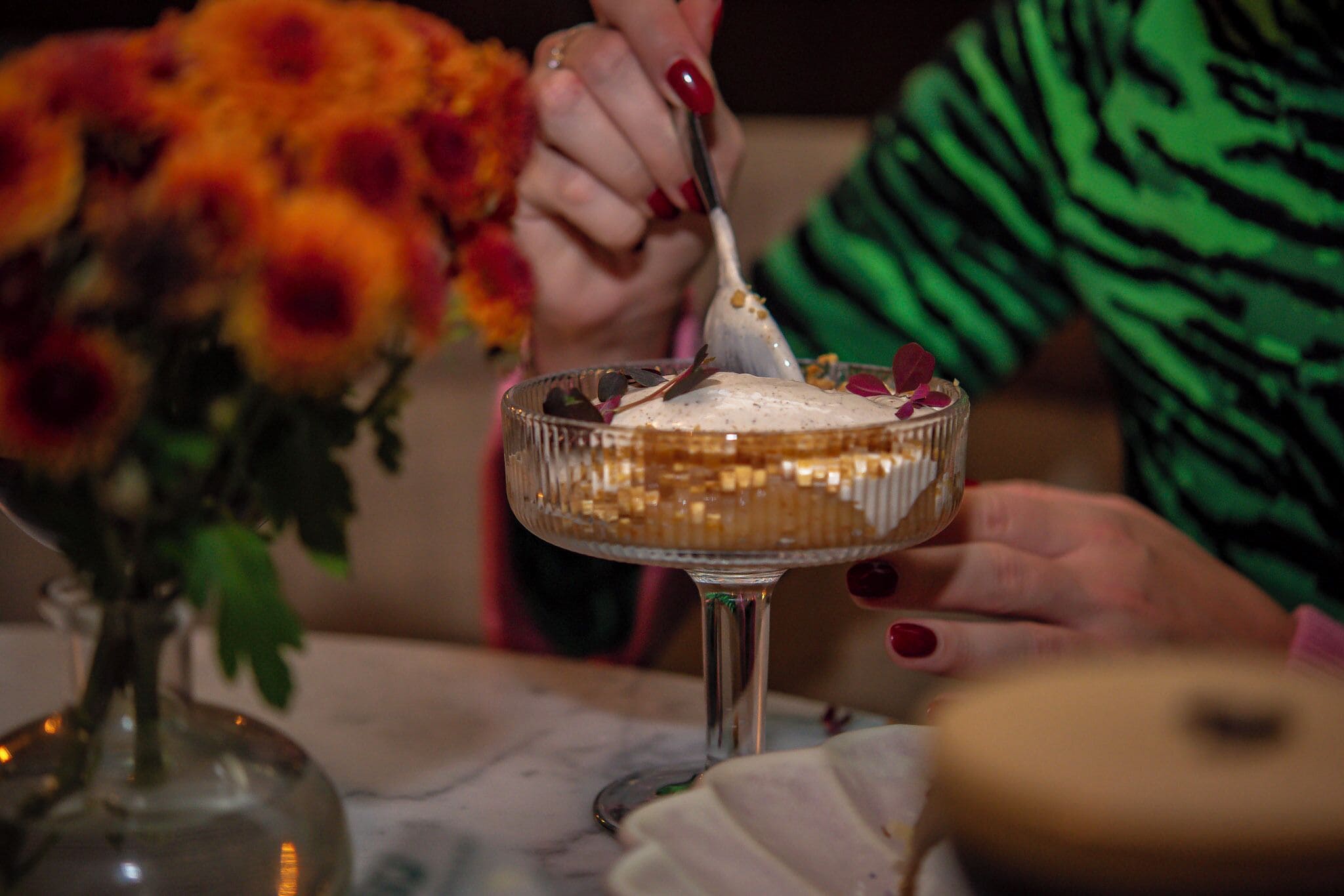 A dessert in a glass bowl with a spoon, a vase with flowers, and a person with red nail polish.