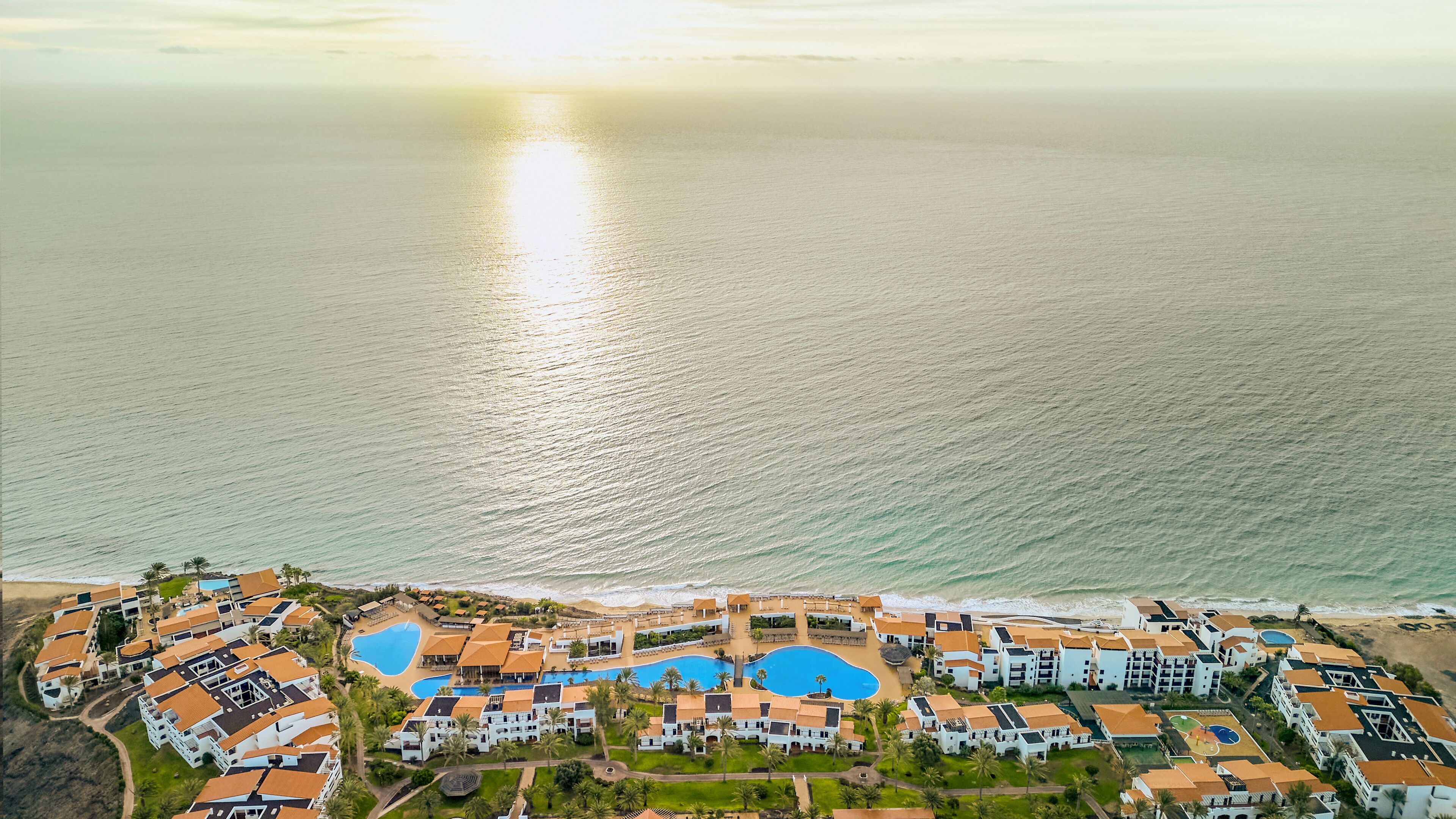 An aerial view of a coastal resort with multiple buildings, a central swimming pool, and a beach.