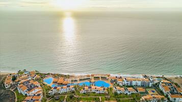 An aerial view of a coastal resort with multiple buildings, a central swimming pool, and a beach.