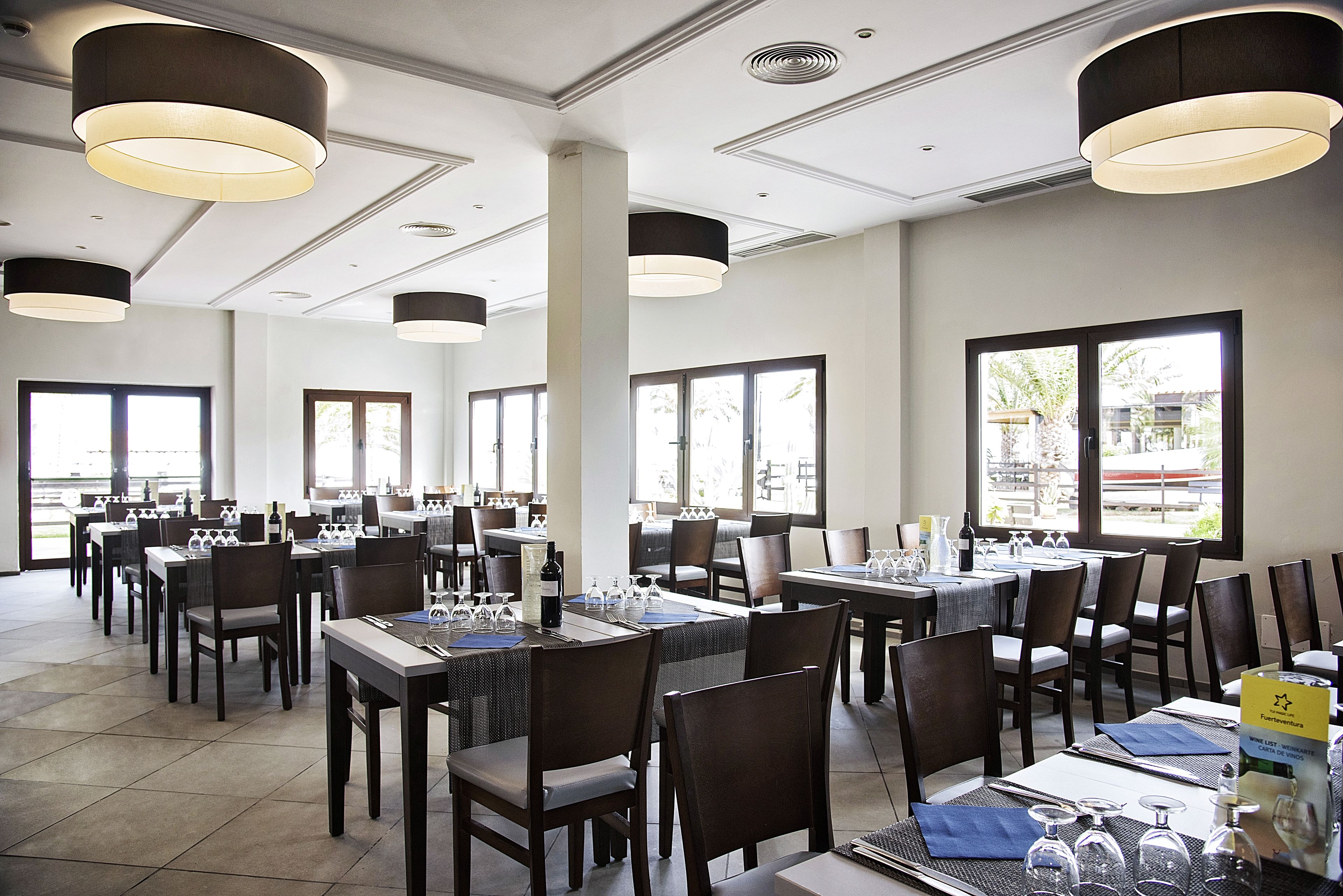 En rummelig restaurant med flere borde og stole, store vinduer og moderne hængelamper.