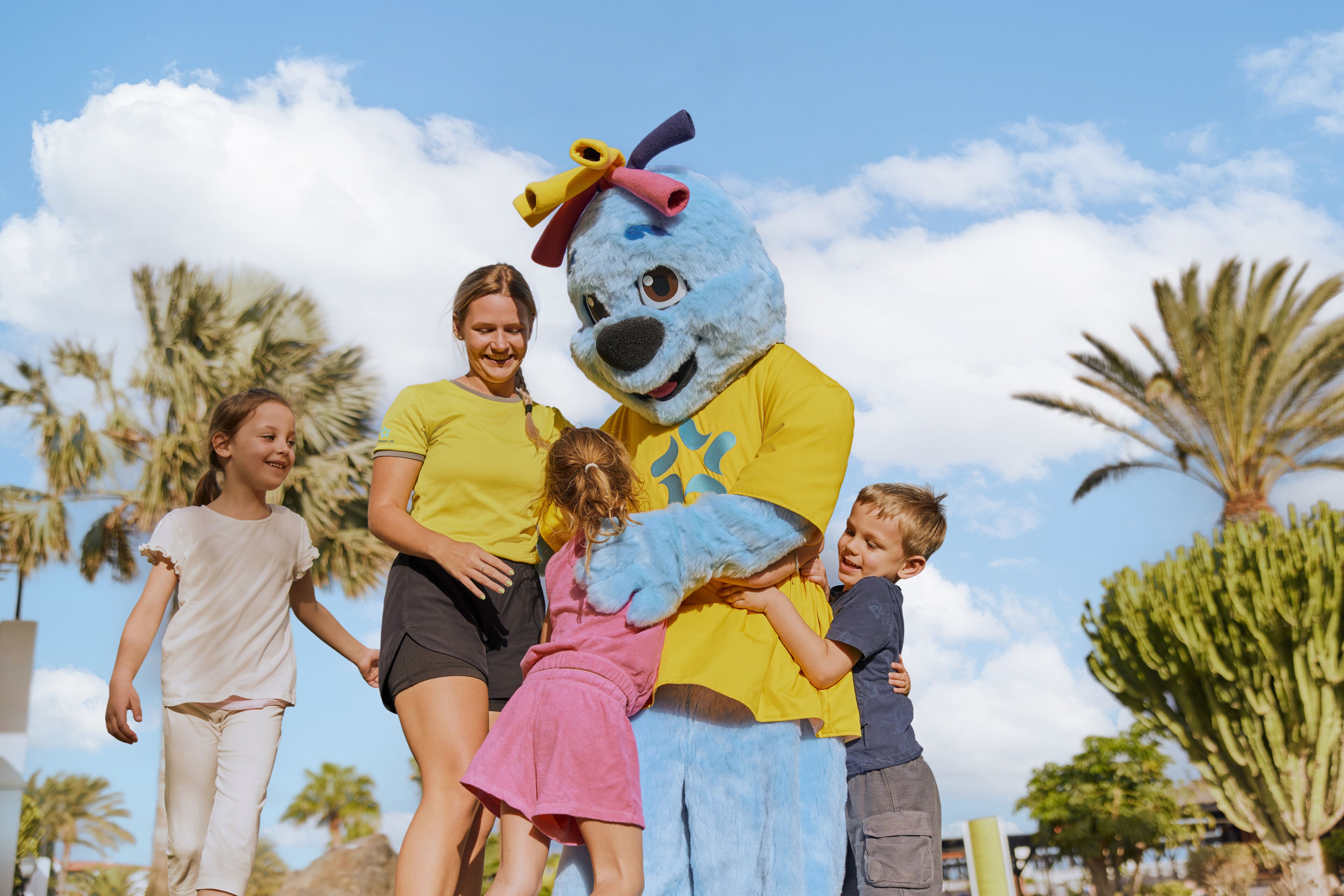 A mascot in a yellow shirt and blue costume interacting with children and adults outdoors.