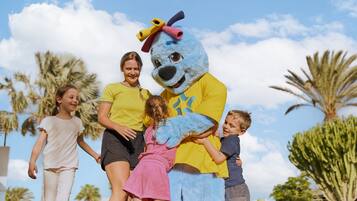 Eine Maskottchenfigur in gelbem Hemd und blauer Kostüm interagiert draußen mit Kindern und Erwachsenen.