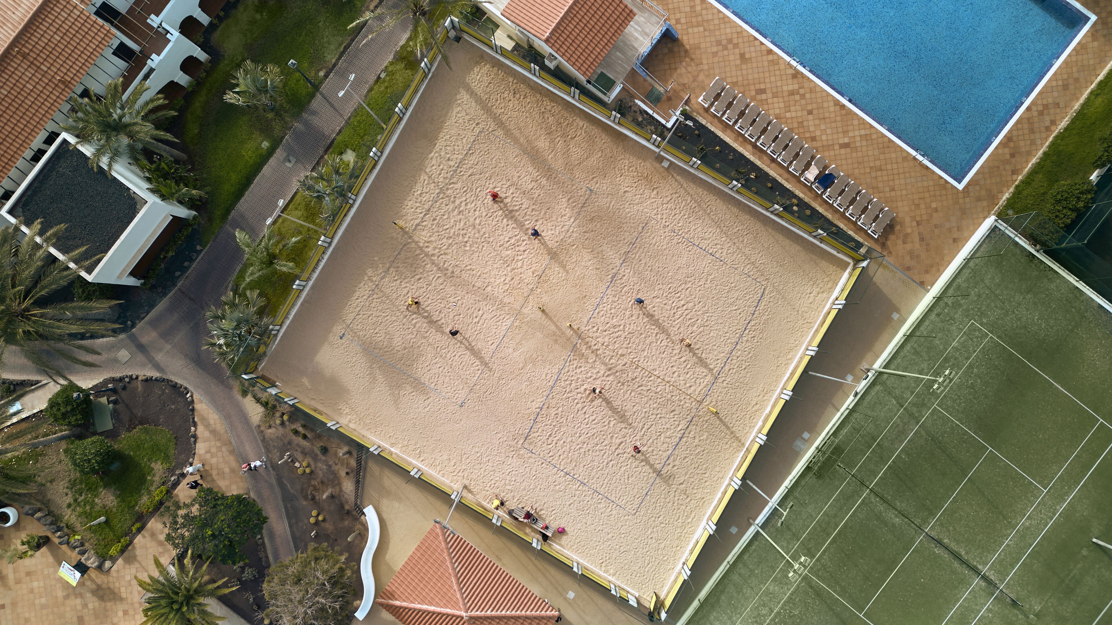 An aerial view of a beach with a volleyball court, a swimming pool, and surrounding buildings.