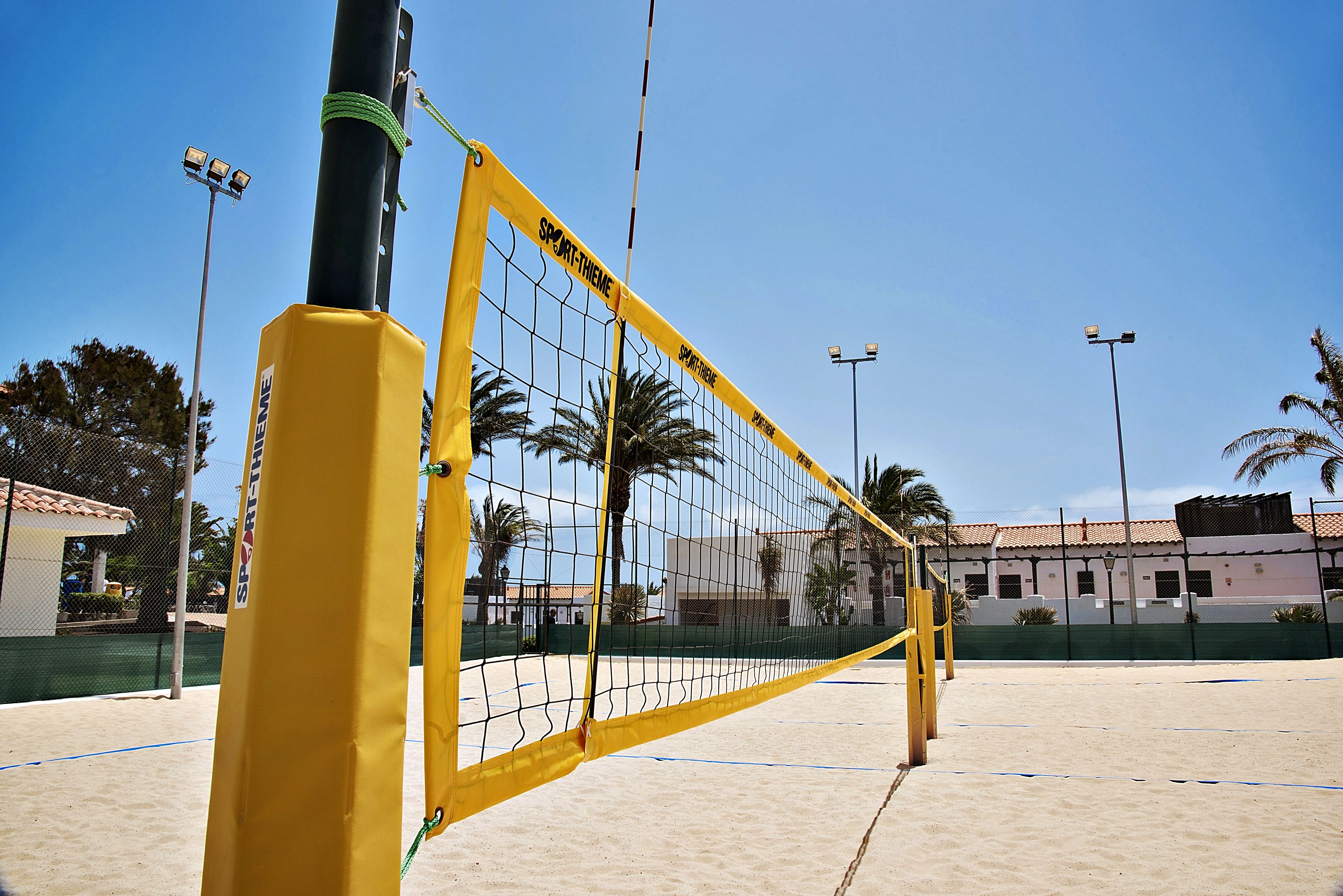 A beach volleyball court with a net, palm trees, and a building in the background.