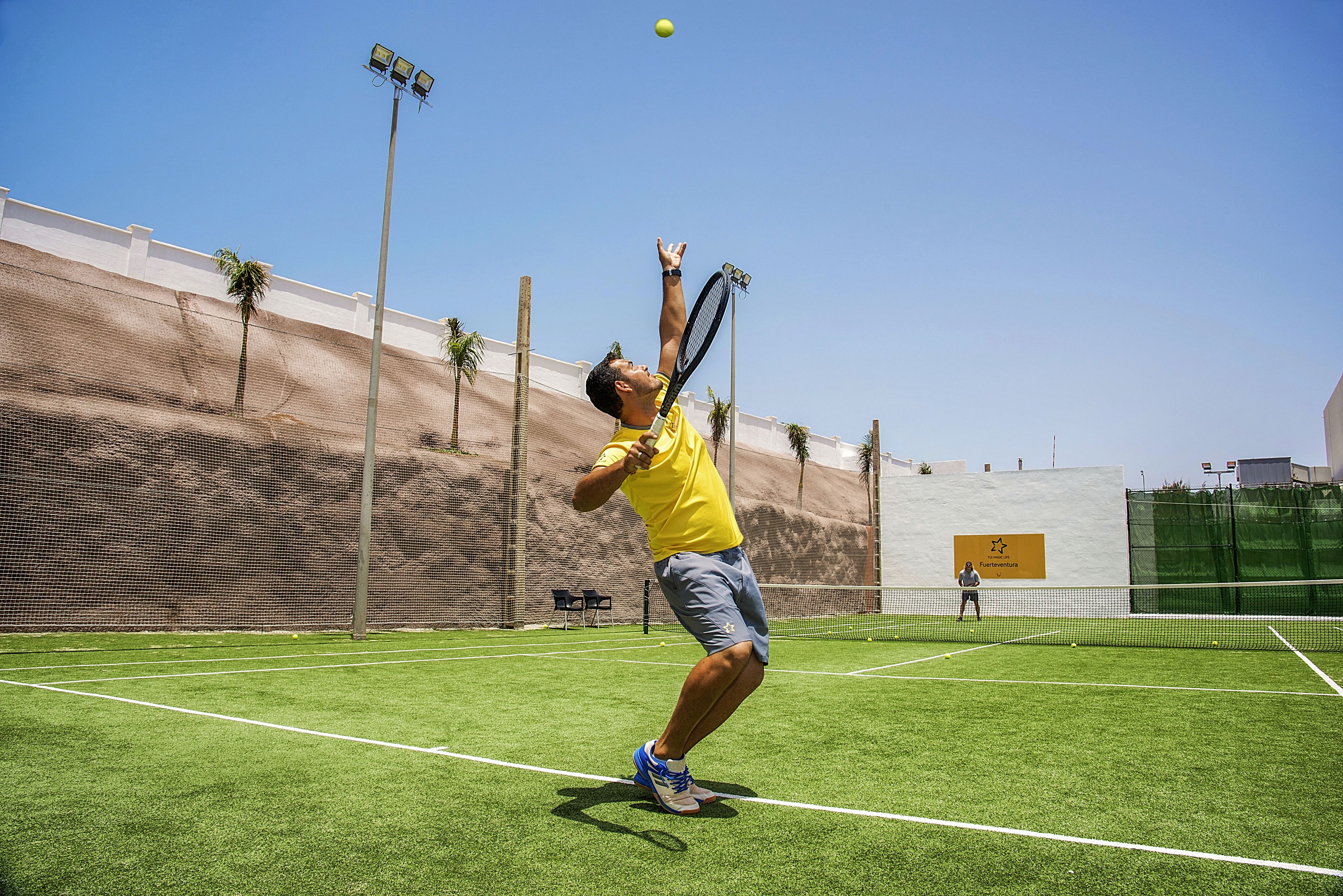 Ein Tennisplatz mit Netz, ein Spieler in einem gelben Shirt und ein klarer Himmel.