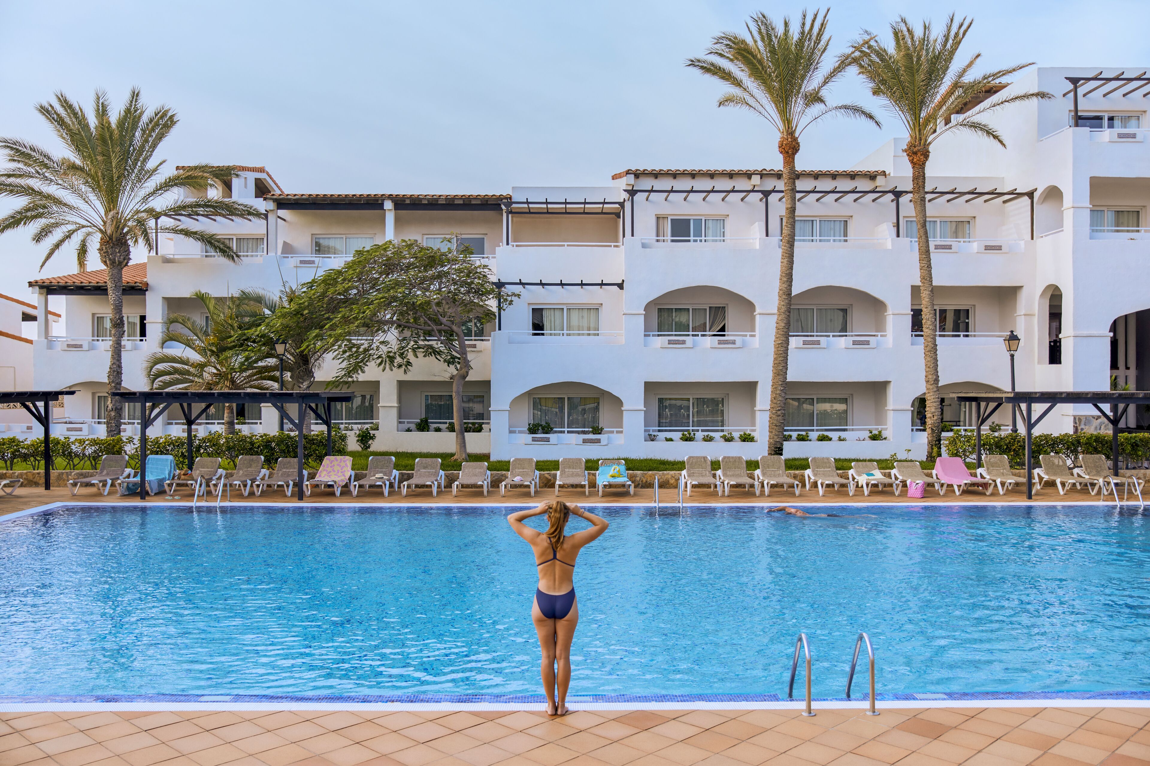 Ein Pool mit Liegestühlen und Palmen, umgeben von einem mehrstöckigen Gebäude mit Balkonen.