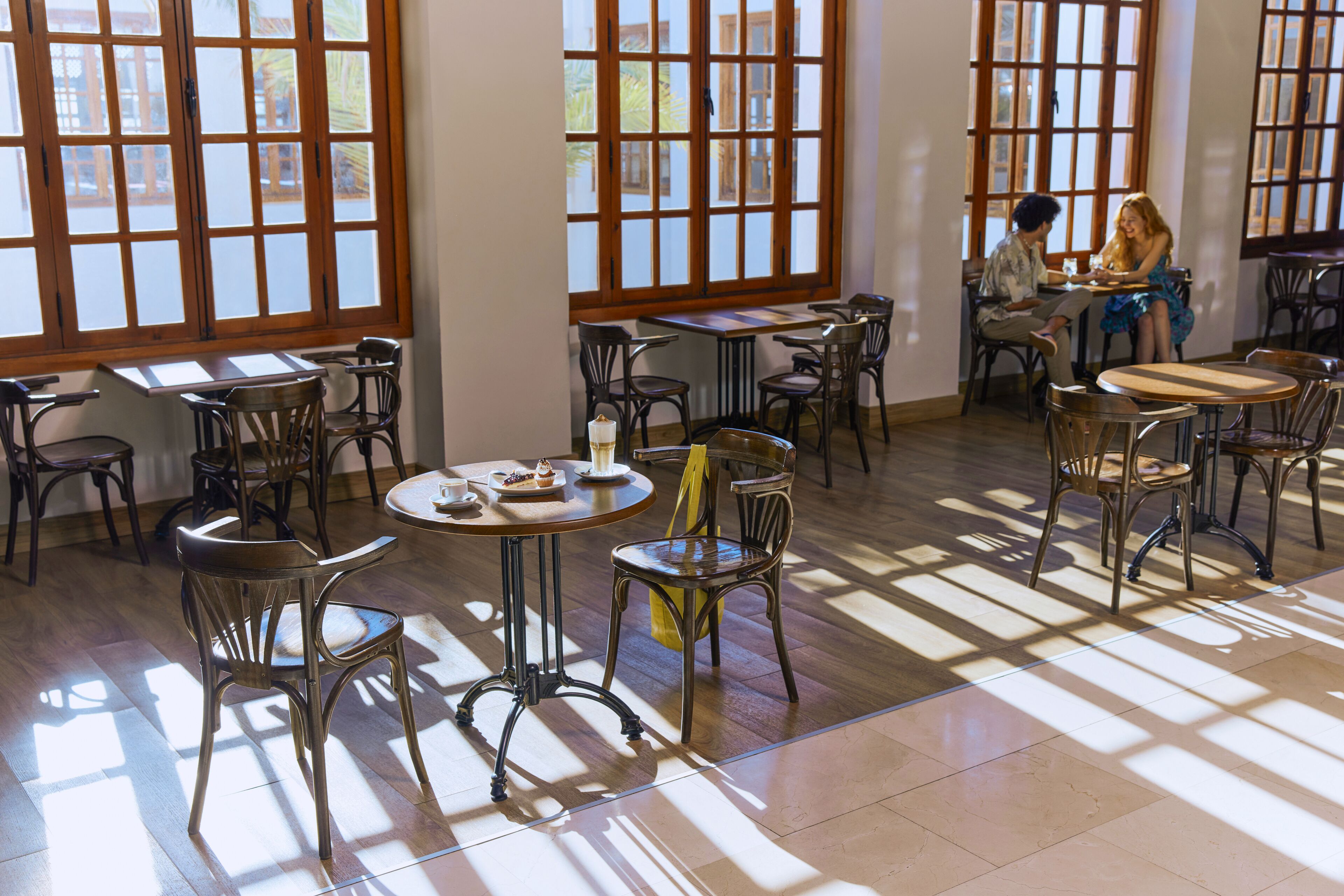 A spacious dining area with multiple tables and chairs, large windows with wooden frames, and a tiled floor.