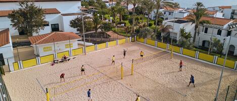 A beach volleyball court with players, surrounded by buildings and palm trees.
