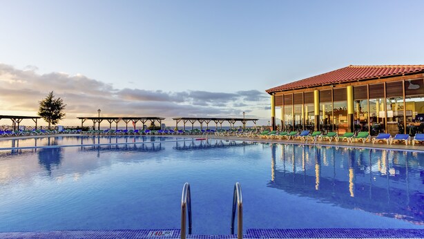 A large outdoor swimming pool with clear blue water, surrounded by lounge chairs and a covered area with large windows. - TUI MAGIC LIFE Fuerteventura (Pajara)