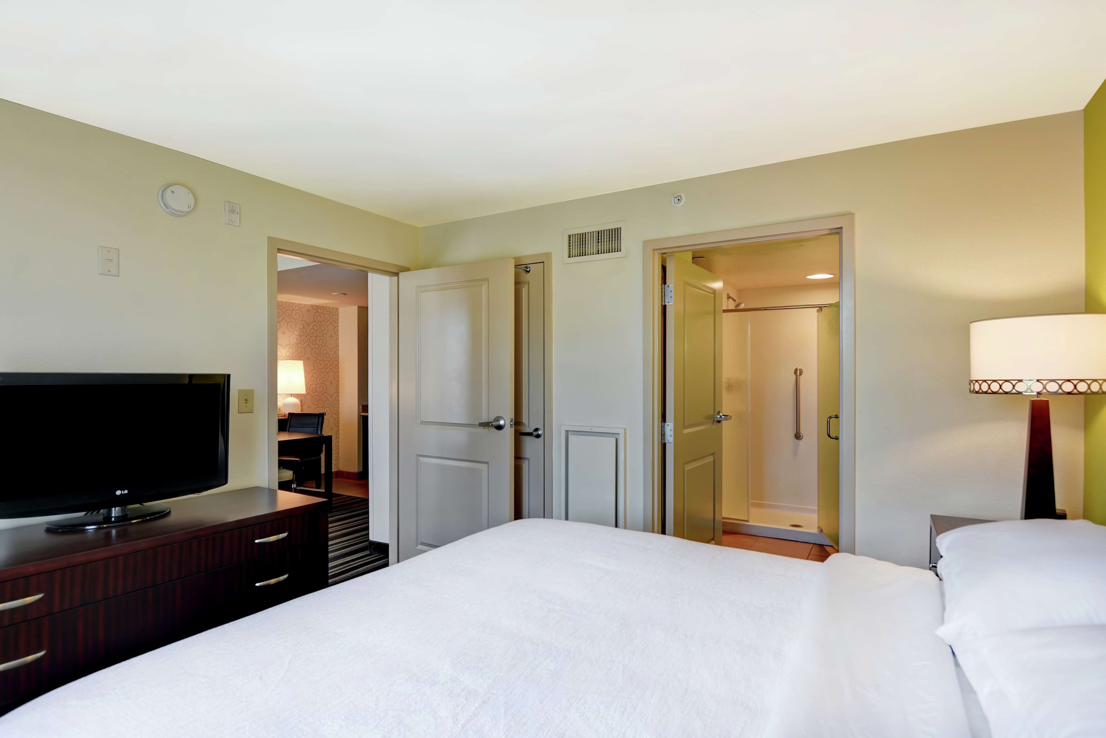 A hotel room with a bed, a TV on a wooden dresser, a lamp, and a door leading to a bathroom.