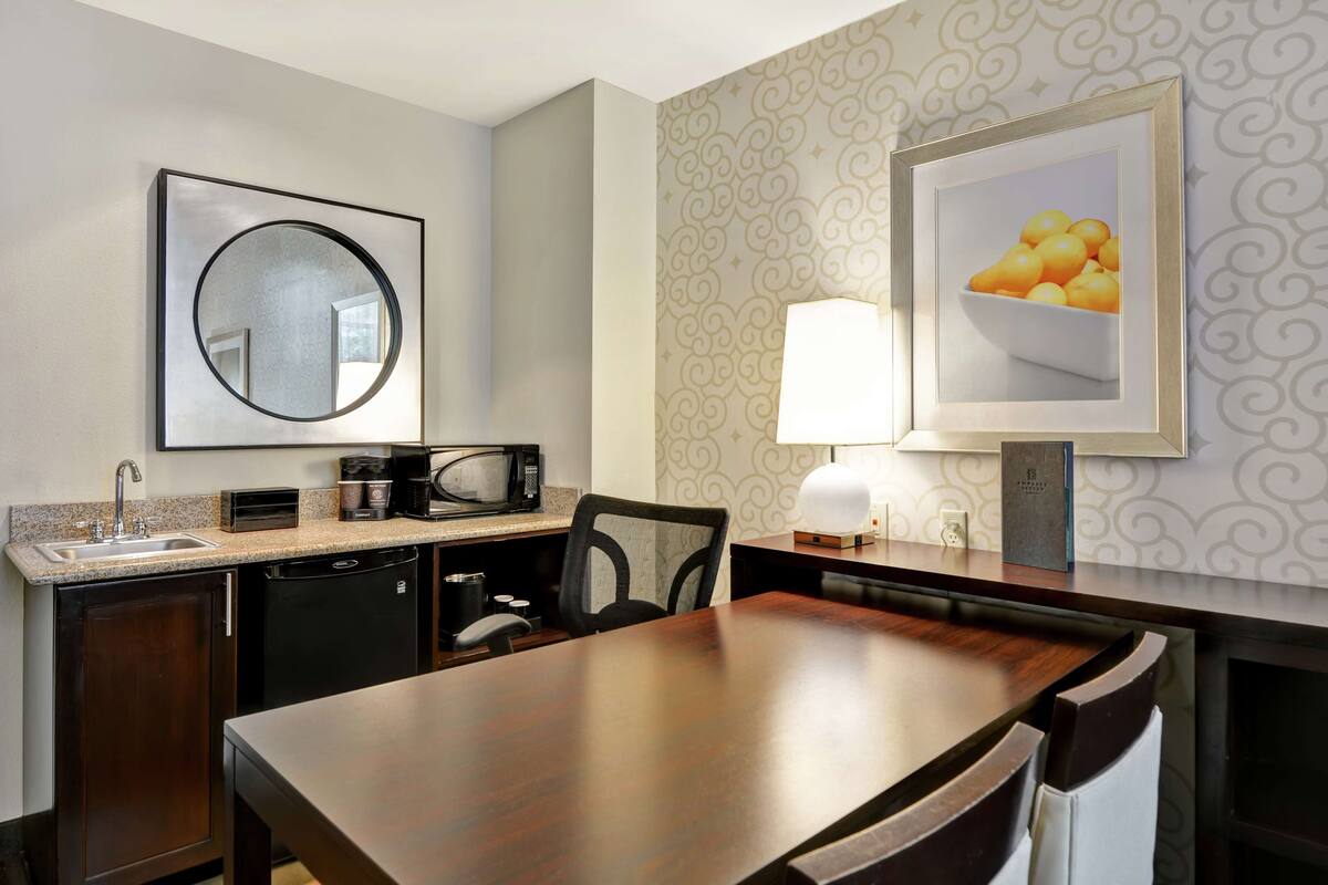 Habitación de hotel compacta con kitchenette, zona de comedor, espejo grande y un cuadro con fruta en la pared.