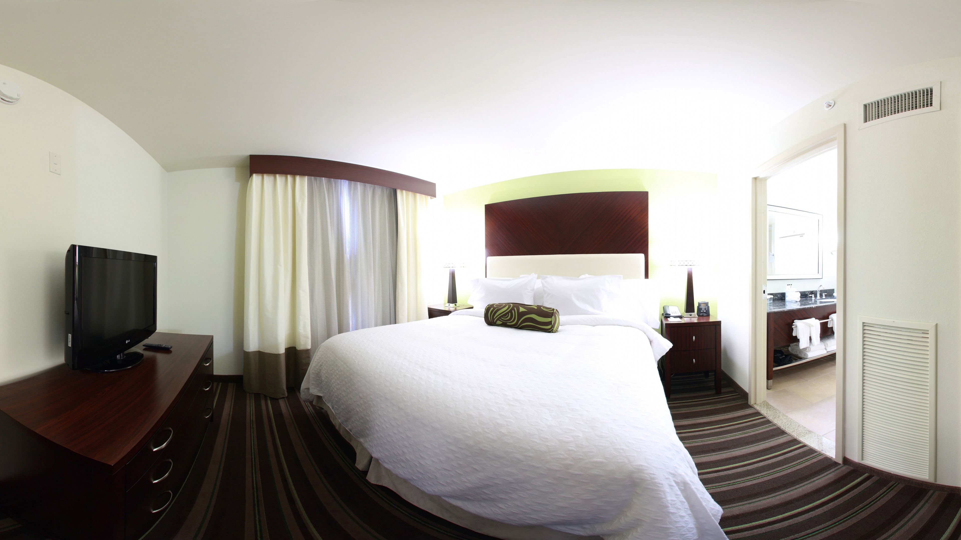 Une chambre d’hôtel avec un grand lit, un téléviseur à écran plat, une table de chevet, une lampe et une salle de bain visible à travers une porte ouverte.