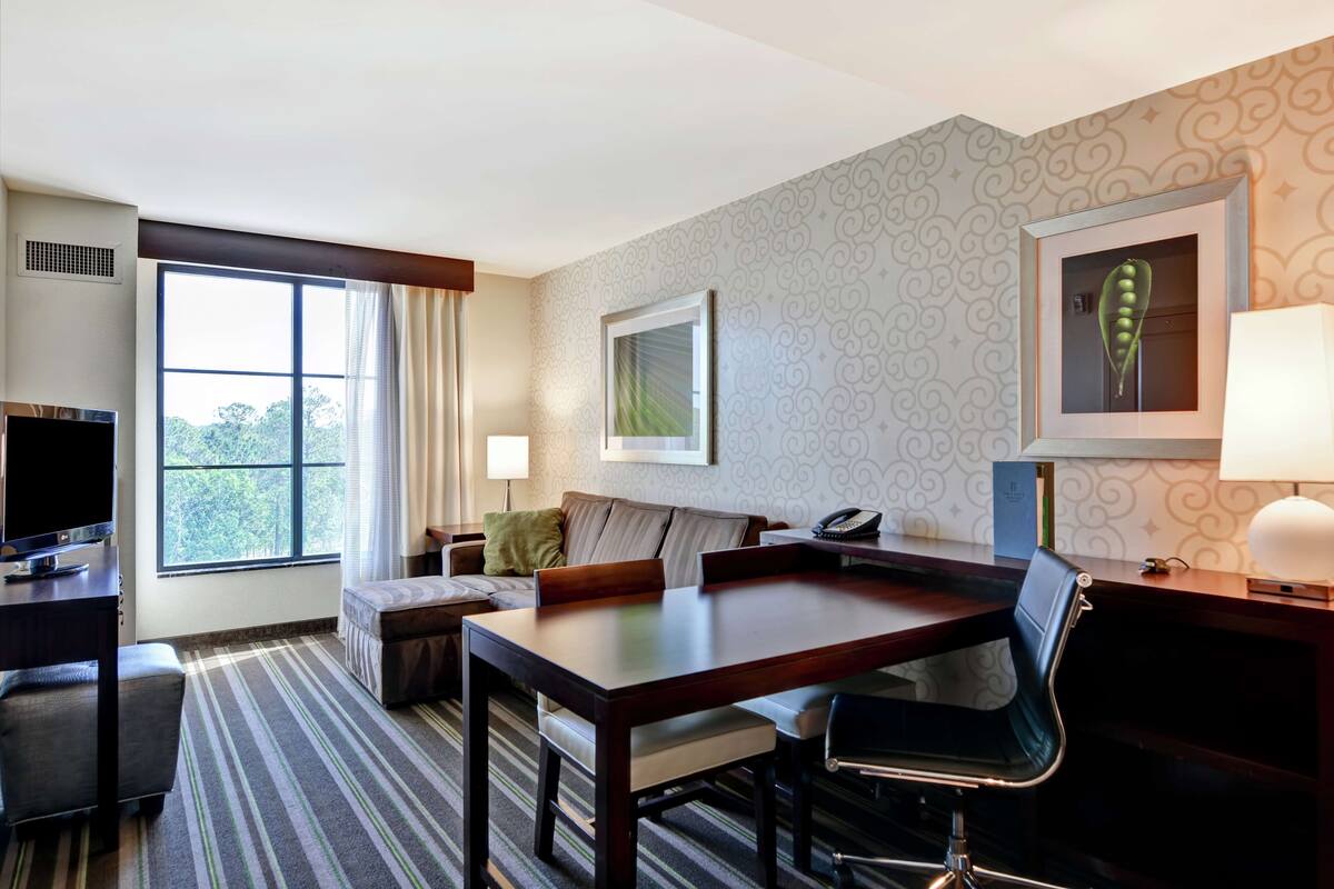 Habitación de hotel con un ventanal grande, un sofá, una mesa de comedor, un televisor y una lámpara.