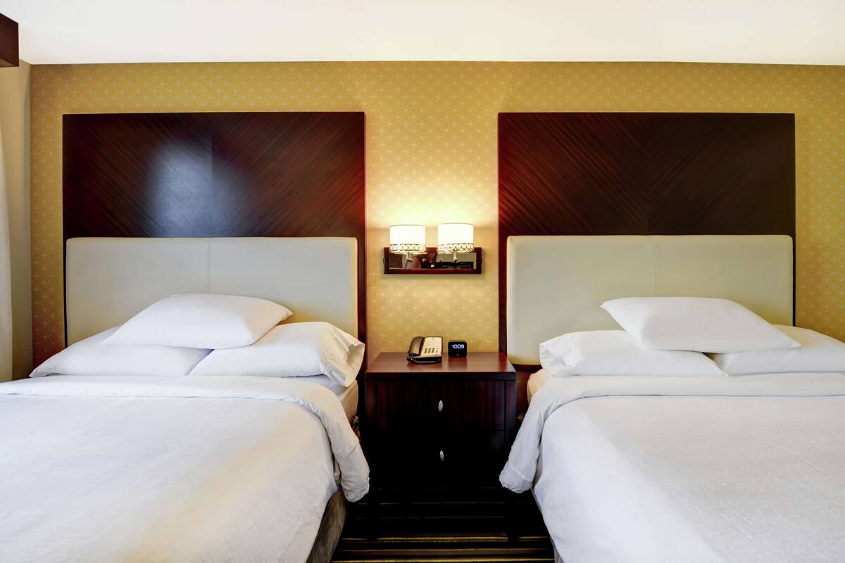 Habitación de hotel con dos camas, una mesita de noche con teléfono y lámparas, y un papel tapiz estampado.