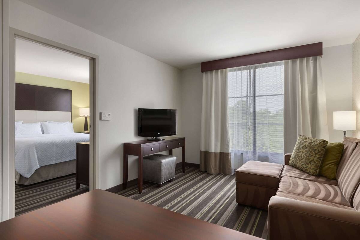 Habitación de hotel con cama, televisor sobre un soporte de madera, sofá y mesa de comedor.