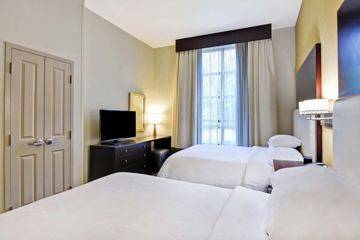 Habitación de hotel con dos camas, un televisor, un escritorio con lámpara y una ventana con cortinas.