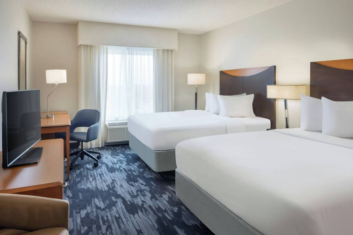 A hotel room with two beds, a desk with a TV, a chair, and a window with curtains.