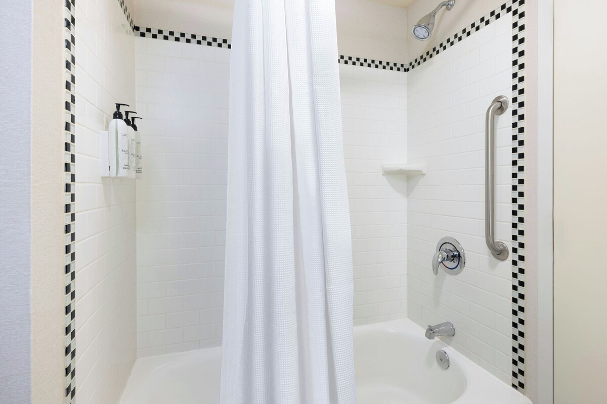 A shower with a white curtain, a handheld showerhead, a handheld showerhead, and a wall-mounted faucet.
