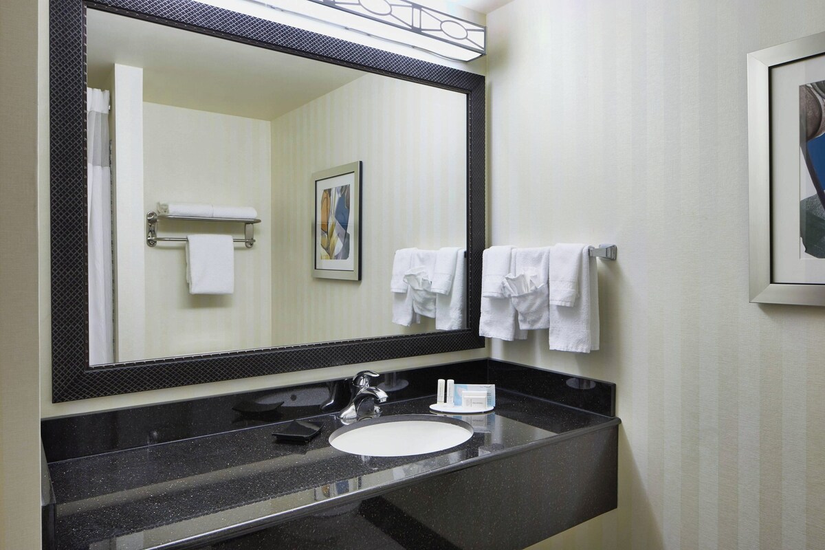 A bathroom with a black countertop, white sink, and a large mirror. There are two white towels hanging on hooks and a framed picture on the wall.