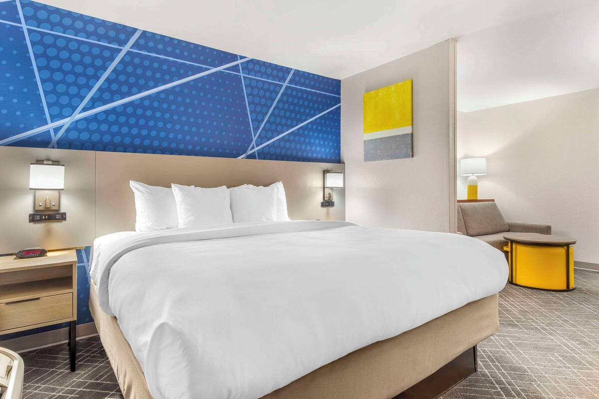 A hotel room with a large bed, a bedside table with a lamp, a chair, and a small table with a lamp.