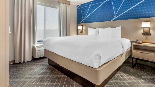 Habitación de hotel con una cama grande, una mesita de noche con lámpara, una ventana con cortinas y una pared con diseño geométrico.