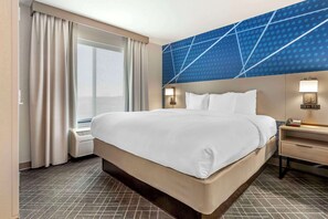A hotel room with a large bed, a nightstand with a lamp, a window with curtains, and a wall with a geometric pattern. - Comfort Suites Whitsett - Greensboro East (Whitsett)