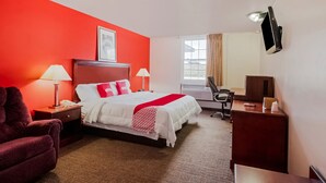 Habitación de hotel con una pared de acento roja, cama con ropa de cama blanca y almohadas rojas, escritorio con silla, televisor montado en la pared y ventana con cortinas.