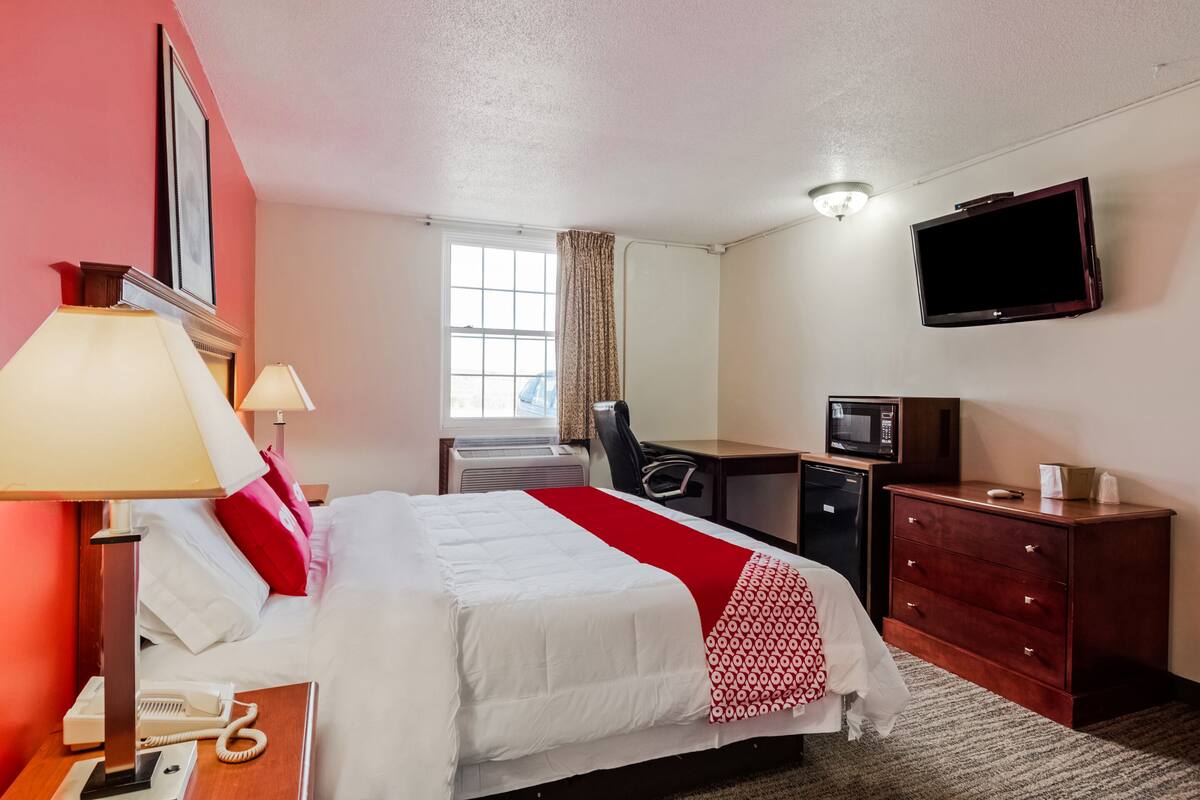 A hotel room with a bed, bedside lamps, a desk with a computer and a TV, a dresser, and a window with curtains.