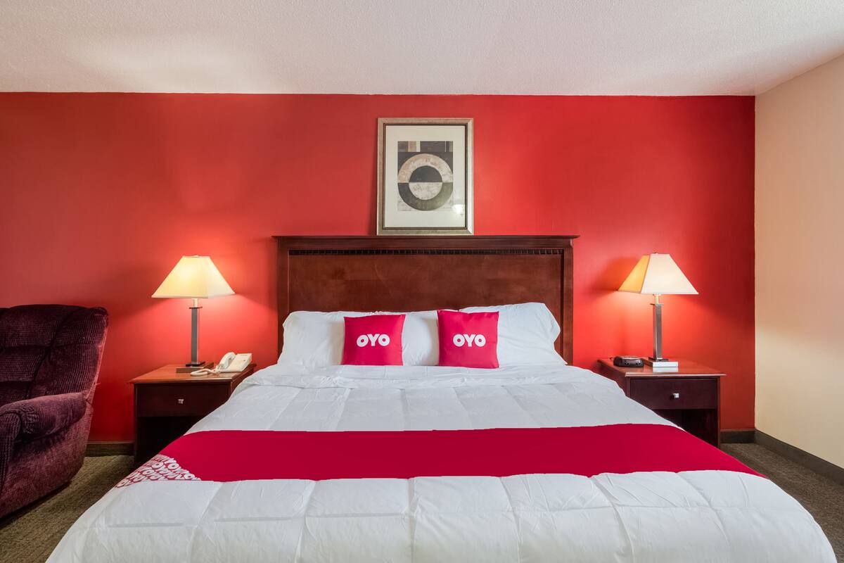 A hotel room with a red wall, a large bed with white bedding, two red pillows, two bedside lamps, and a framed artwork above the bed.