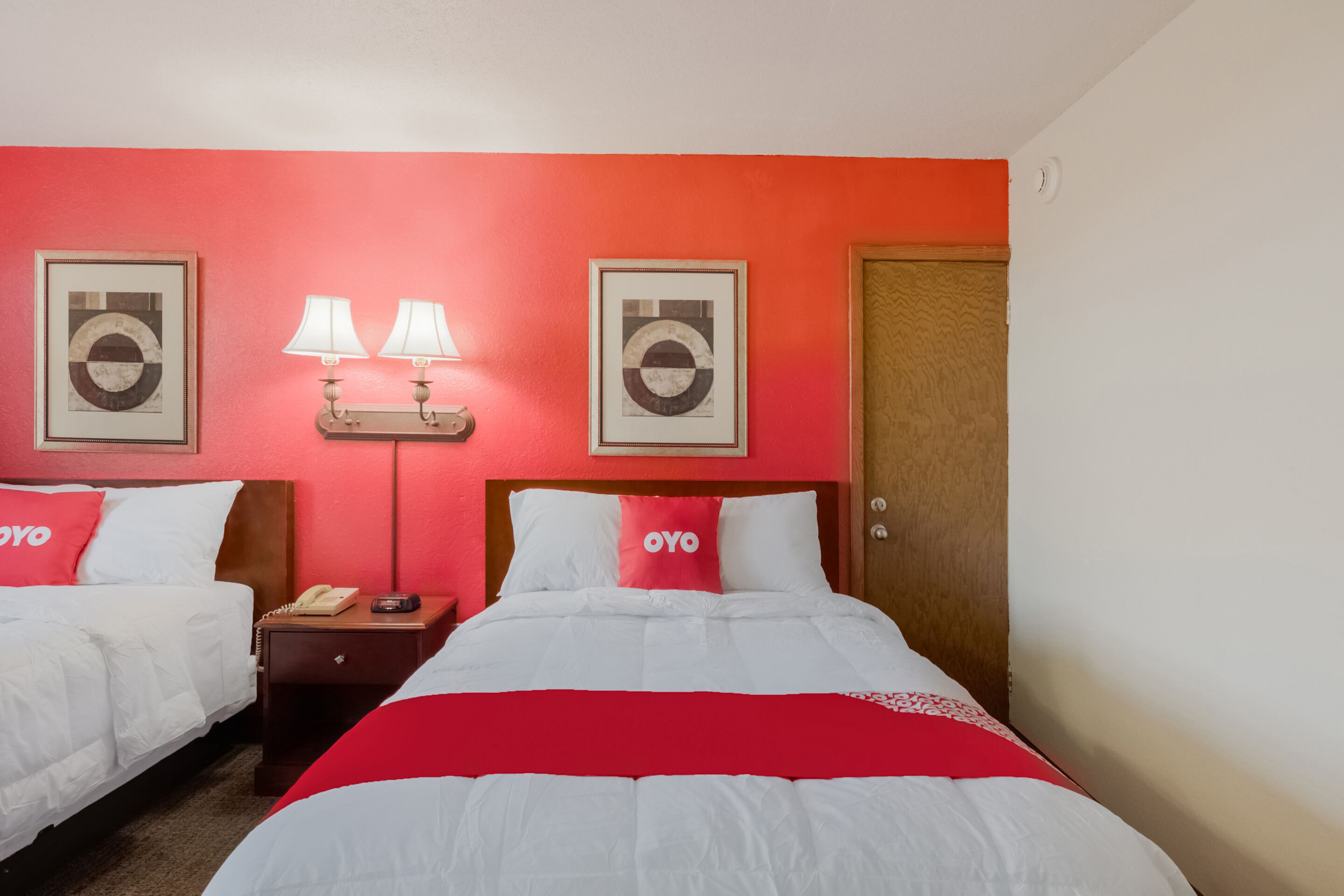 Habitación de hotel con dos camas, un cabecero de madera, una pared roja, dos cuadros enmarcados, una mesita de noche con una lámpara y un teléfono.