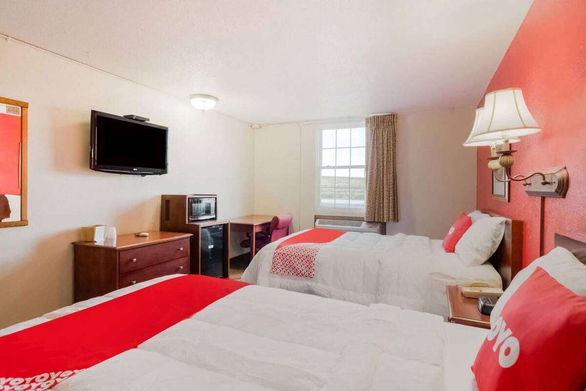 A hotel room with two beds, a TV, a desk with a computer, a window with curtains, and a lamp.