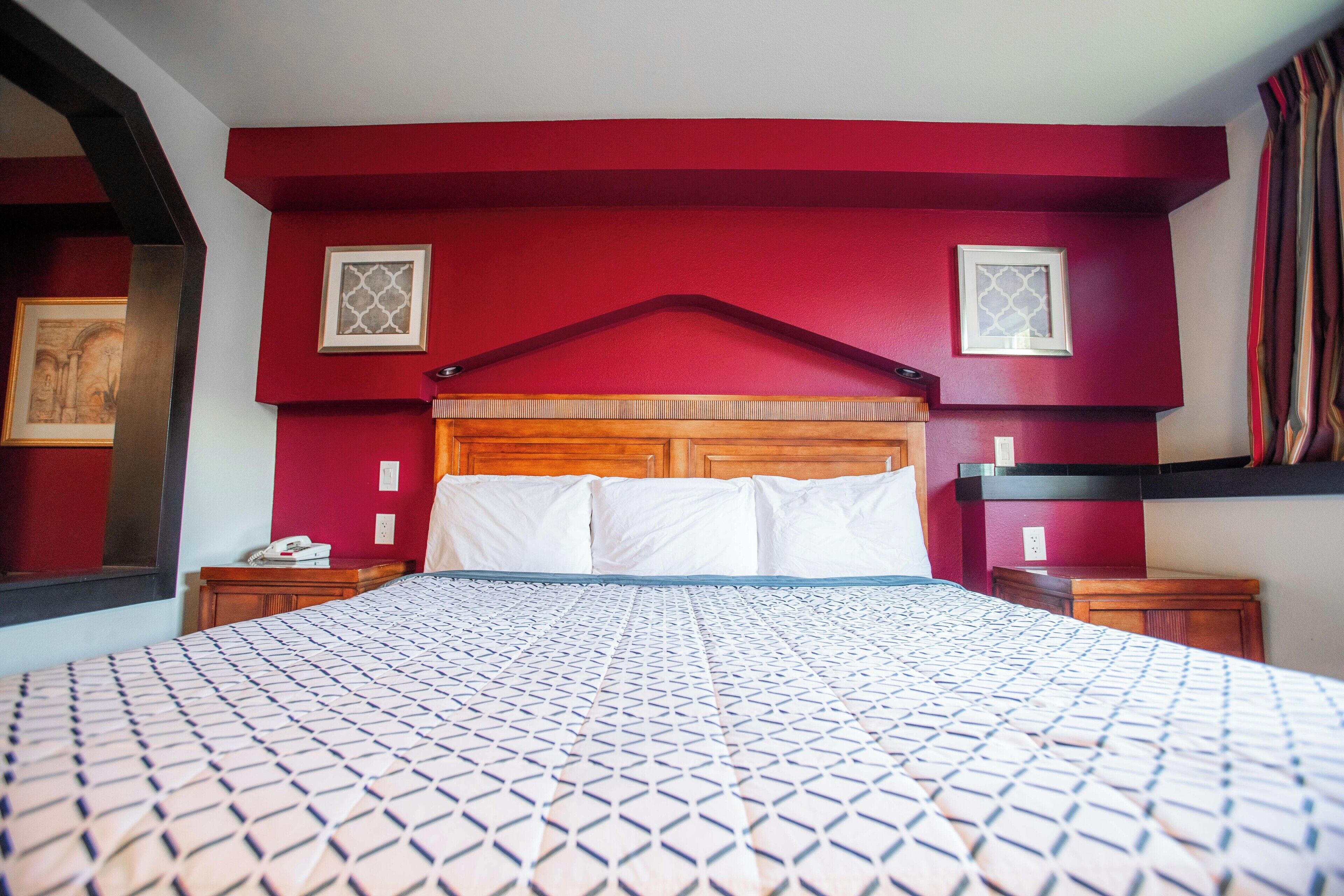 Quarto de hotel com cama grande, cabeceira de madeira, paredes com detalhes vermelhos e duas mesas de cabeceira com luminárias.