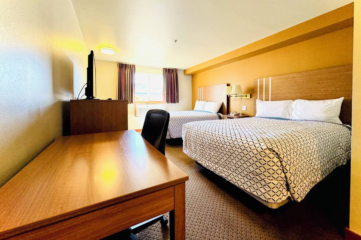A hotel room with a wooden desk, two beds with patterned bedding, a nightstand with a lamp, and a window with curtains.