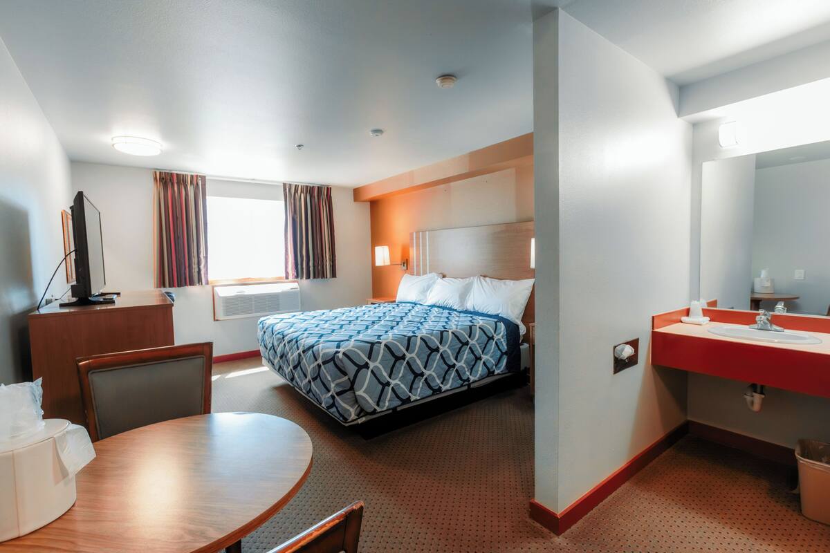 A hotel room with a bed, a desk, a chair, a table, a sink, and a mirror.
