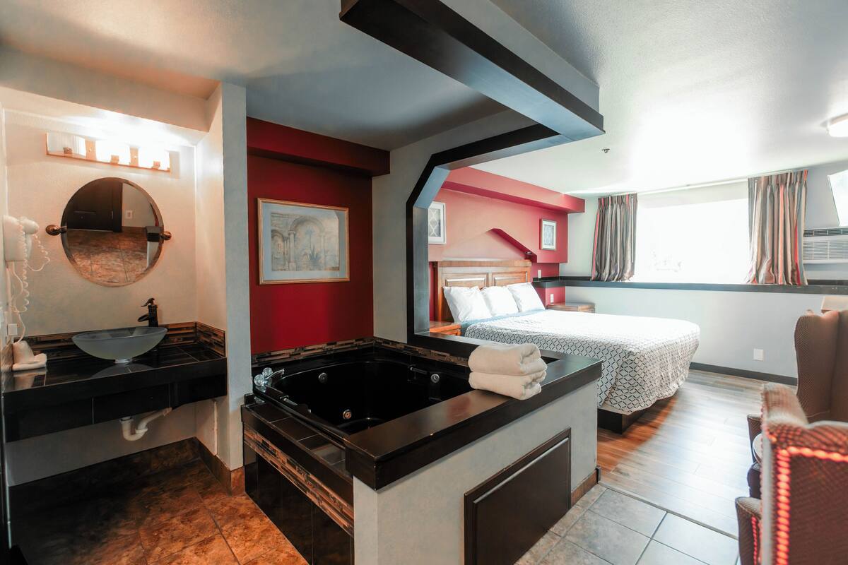 A hotel room with a black bathtub, a bed with a patterned bedspread, a round mirror, and a red accent wall.