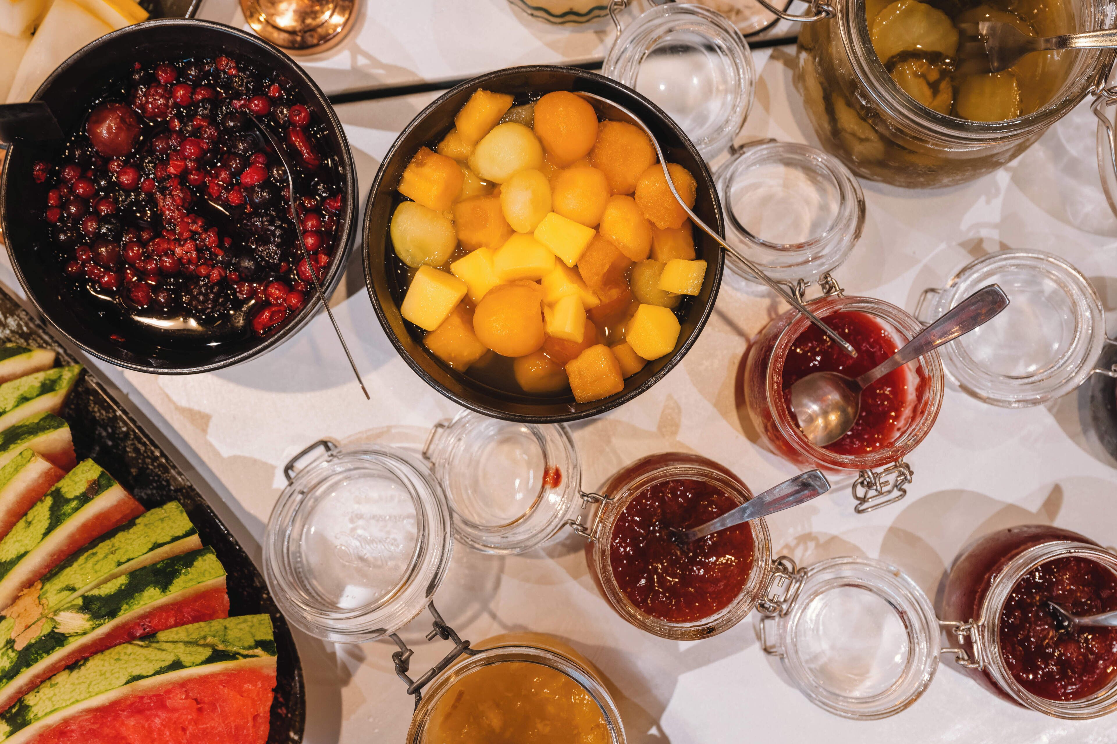 A table with various food items and condiments, including a bowl of diced fruit, a bowl of berries, and several jars of preserves.