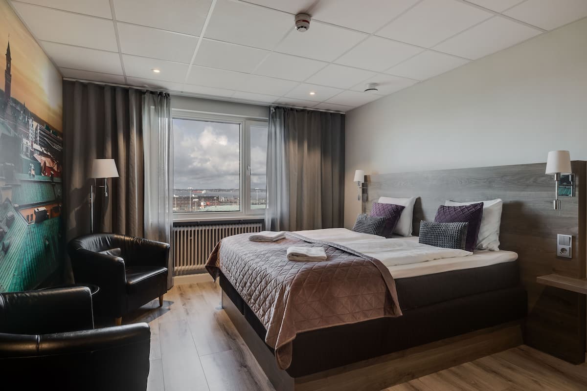 Una habitación de hotel moderna con una cama grande, vistas a la ciudad, un rincón de descanso con una silla y un cuadro en la pared.