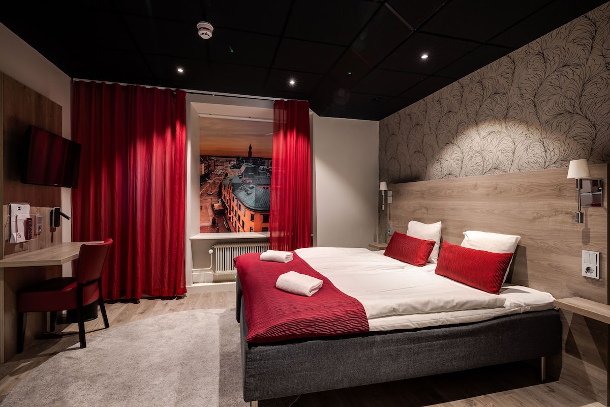 Una habitación de hotel moderna con una cama grande, cortinas rojas, un escritorio y vistas a la ciudad a través de una ventana.