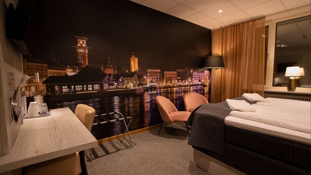 Habitación de hotel con cama, escritorio, silla y vista a un paisaje urbano nocturno.