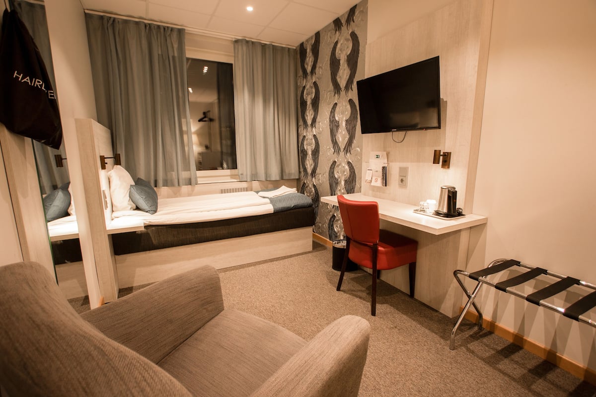 Habitación de hotel con cama, escritorio, silla, televisor y ventana con cortinas.
