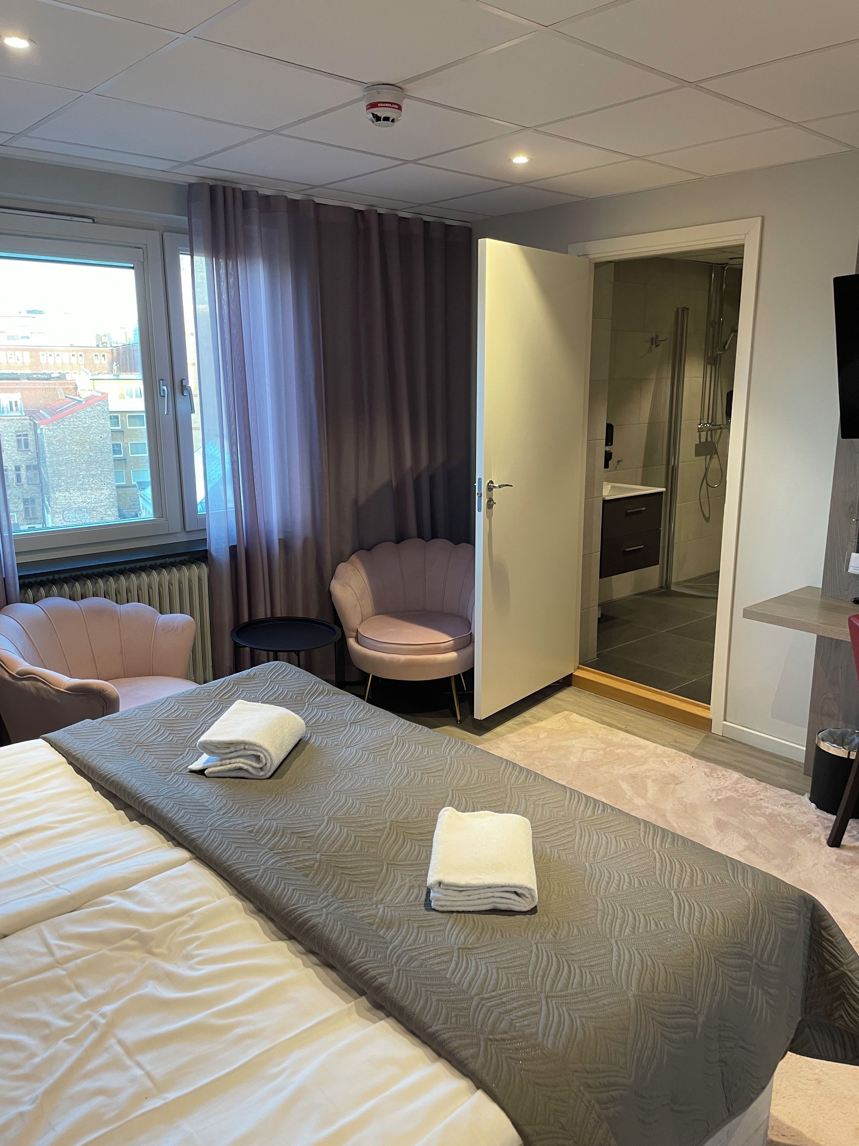 A hotel room with a bed, a chair, a bathroom with a shower, and a view of buildings outside.